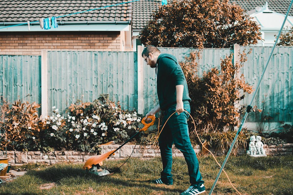 A man wearing glasses, a dark green hoodie, and jeans is using an orange electric lawn mower to trim a well-maintained grassy area of a garden. The garden features a neatly edged lawn, flower beds wit
